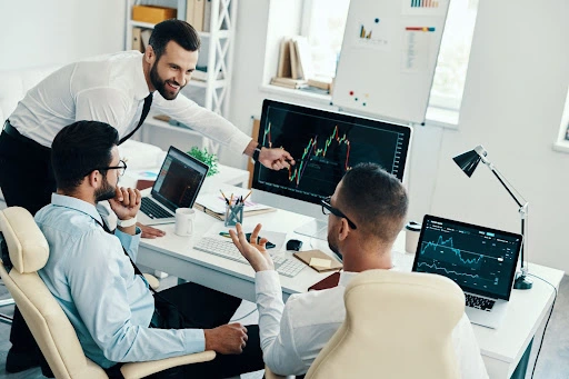 three businessmen engaging in a discussion regarding the stocks market graph shown on the monitor screen, and two laptops